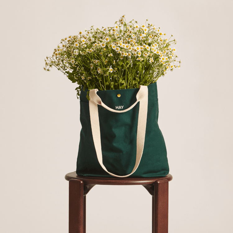 Green organic weekend tote bag filled with white chamomile flowers on a stool. HAY everyday bag, sustainable.