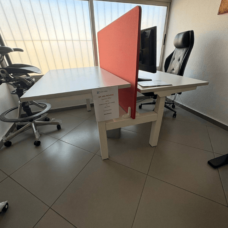 A shared white desk with a red partition, monitor, and black office chair is in a tiled office space.