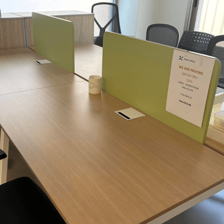 Office desks with light wood grain tops and green dividers, featuring chairs and a promotional sign.