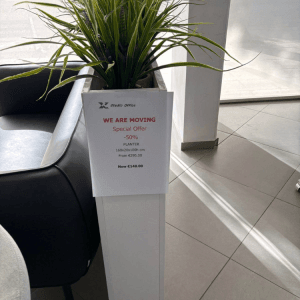 A tall, white planter with green plants sits next to a black couch, with a promotional sign attached.