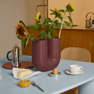 A maroon U-shaped ceramic vase holding sunflowers sits on a light blue table set for breakfast.