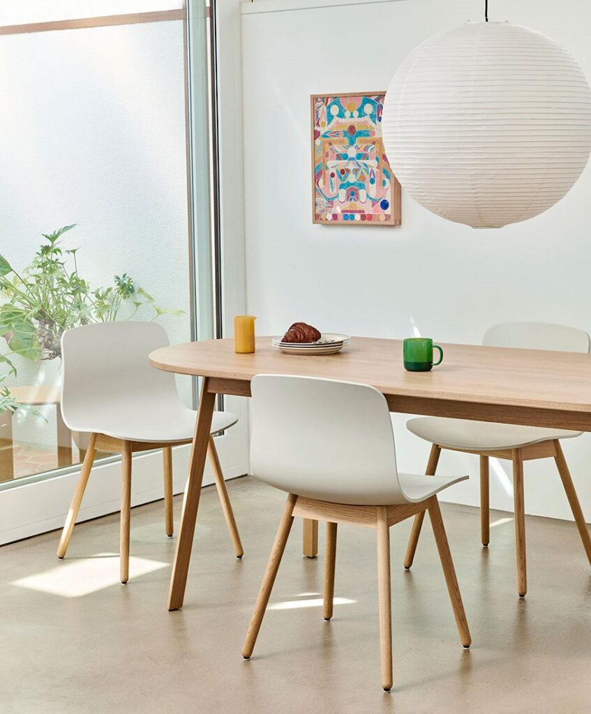 Bright dining area with a light wood table, white chairs, a large paper lantern, and a colorful framed artwork.
