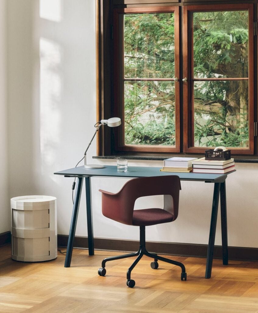 A minimalist home office with a blue desk, maroon swivel chair, books, lamp, and a window overlooking trees.