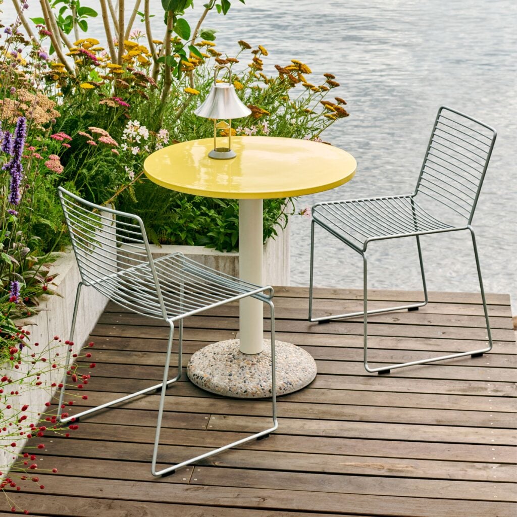 Yellow round outdoor table with two metal wire chairs on a wooden deck beside flowers and water.
