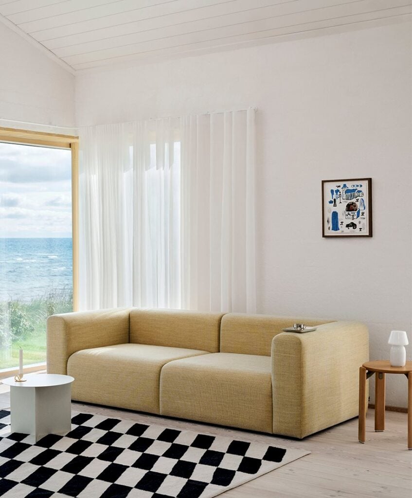 A beige sofa in a bright room with sheer curtains, a checkered rug, and a view of the sea through a large window.