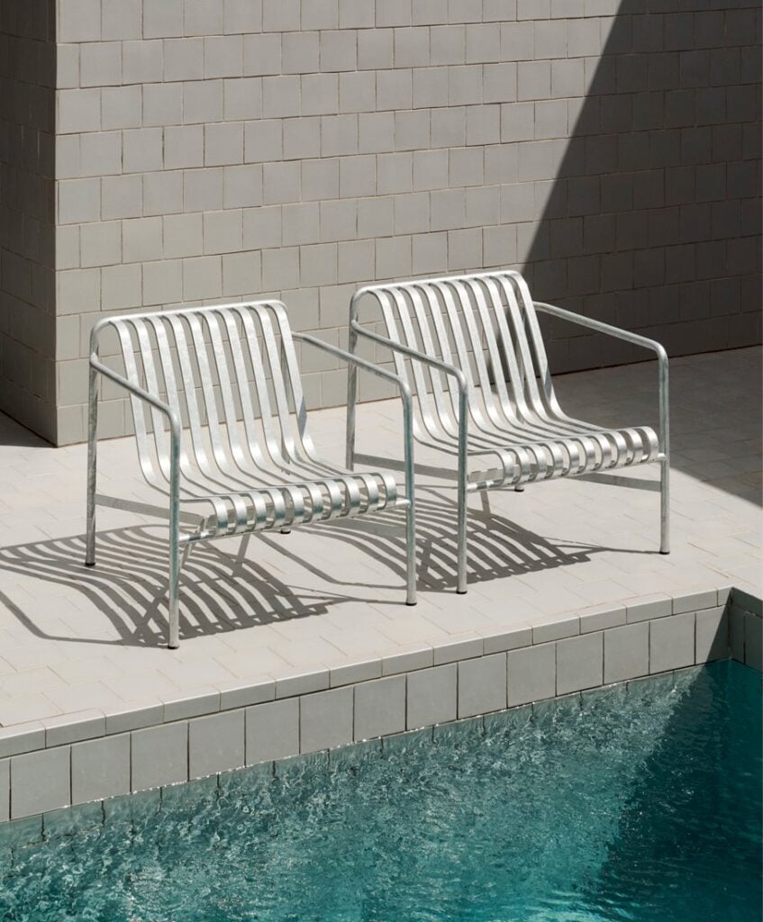Two white metal slatted chairs on a tiled pool deck beside a swimming pool, casting shadows on the tiles.