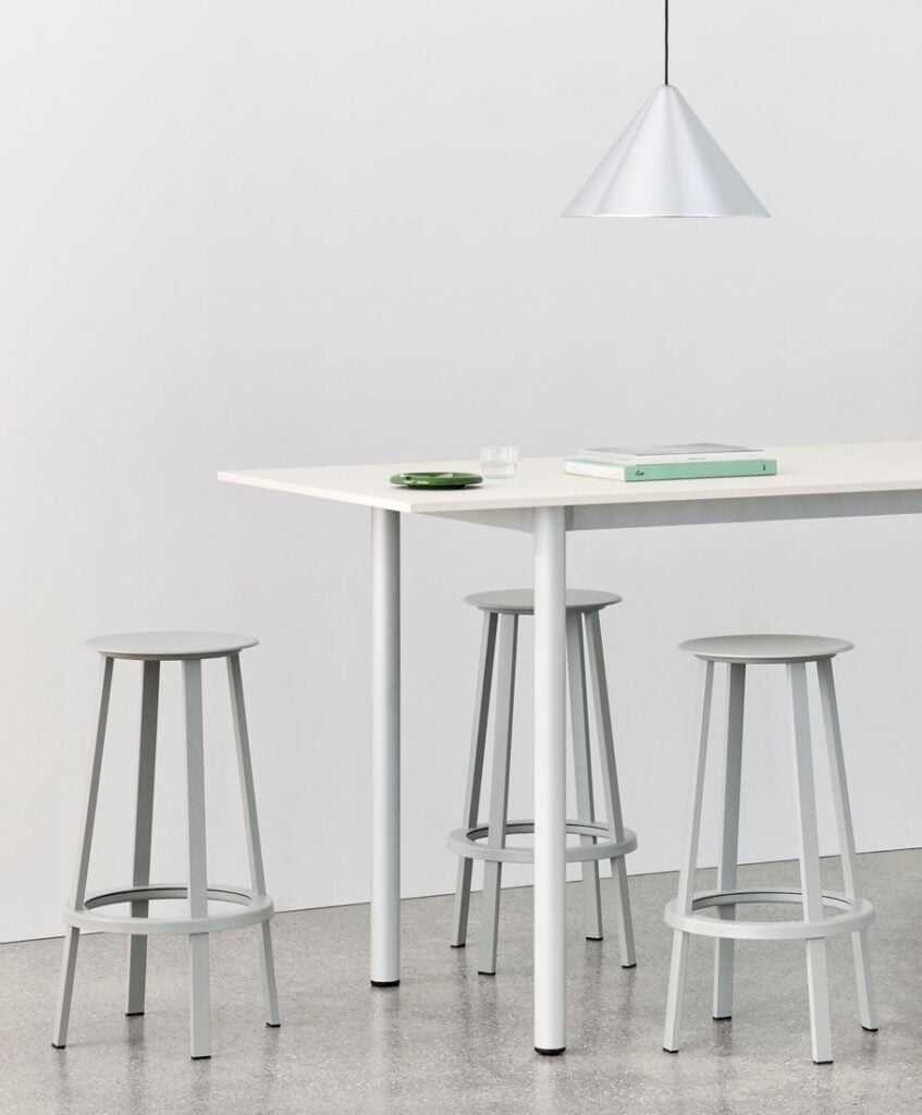 A white table with three grey metal bar stools, a silver cone pendant light above, and a book and bowl on top.