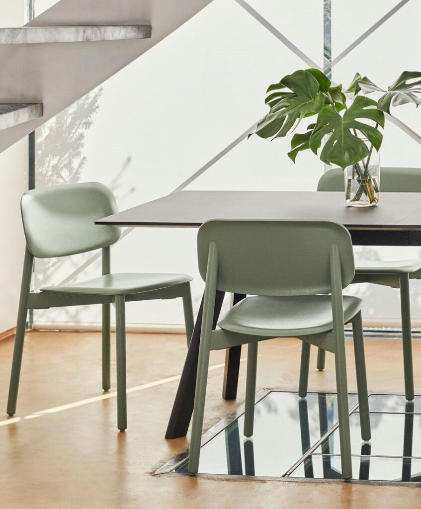 Green molded plastic chairs with wooden legs arranged around a dark table with a glass vase holding monstera leaves.